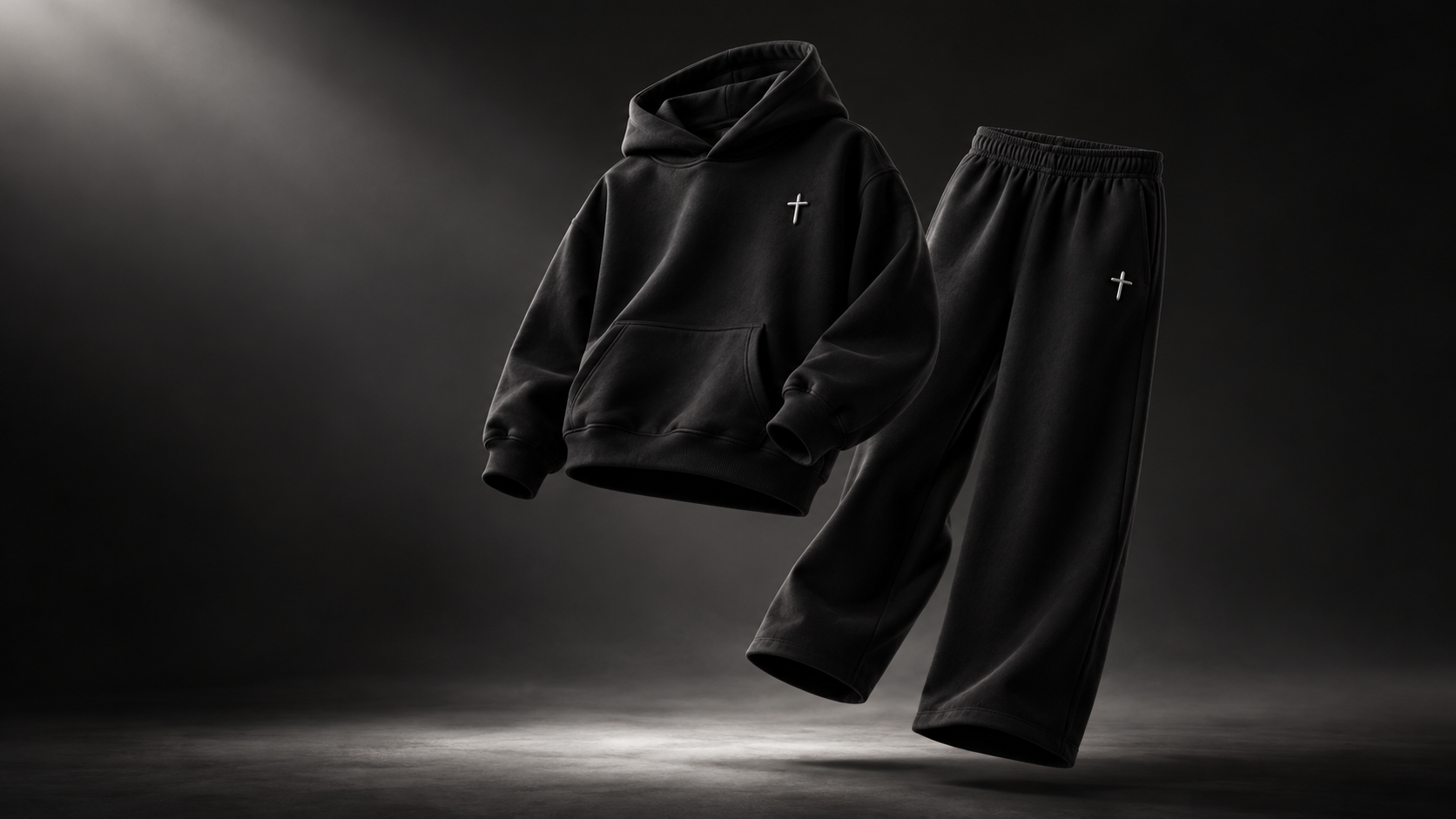 Editorial product concept image of a floating black VIA hoodie and matching wide-leg pants with small silver cross emblems